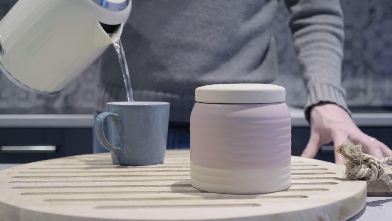 Man pours hot water into mug for tea as camera slides forward slowly. Mug sits on wooden display board with a ceramic jar.