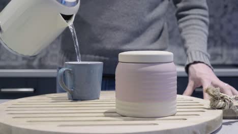 man pours hot water into mug for tea as camera slides forward slowly. mug sits on wooden display board with a ceramic jar.