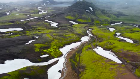 aerial drone flying over green mossy and dark rocky volcanic iceland terrain landscape with white snow on it. large mountains with steep cliffs and edges visible in the background. cloudy weather.
