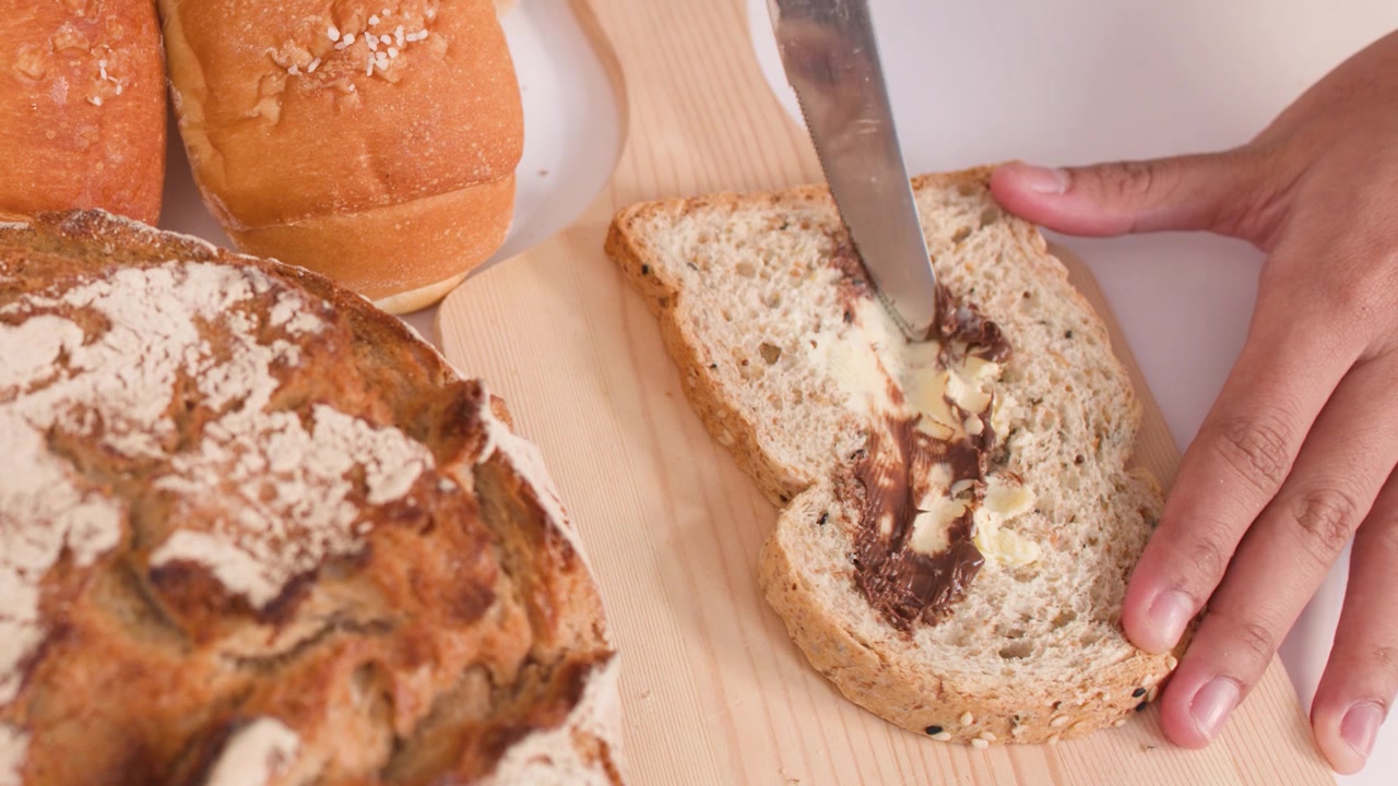Overhead shot of hands spreading peanut butter and butter onto multigrain bread with a knife.
