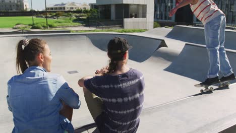 back view of caucasian woman and man sitting, looking at skateboarding friend on sunny day