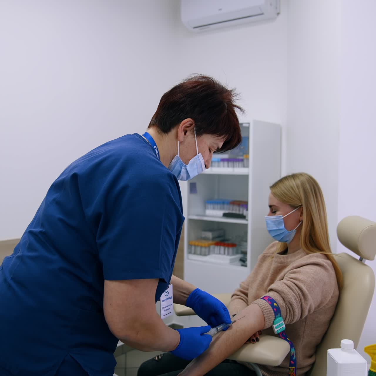 Female nurse takes the blood from patient's vein. Blonde woman in mask doesn't look at the syringe filling with her blood