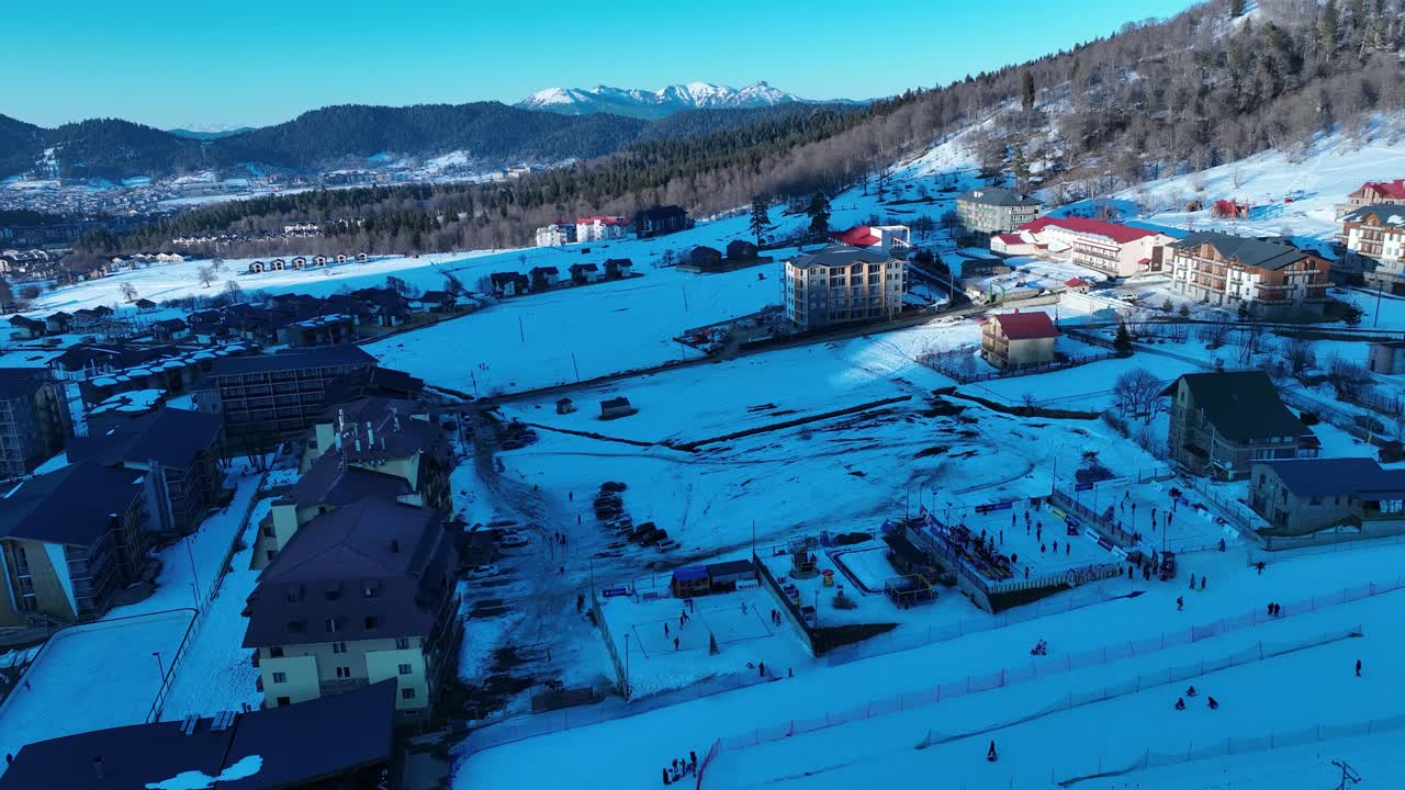An aerial view of a snow-covered mountain resort at dusk, with lights illuminating the lodges and facilities, creating a warm and inviting winter scene