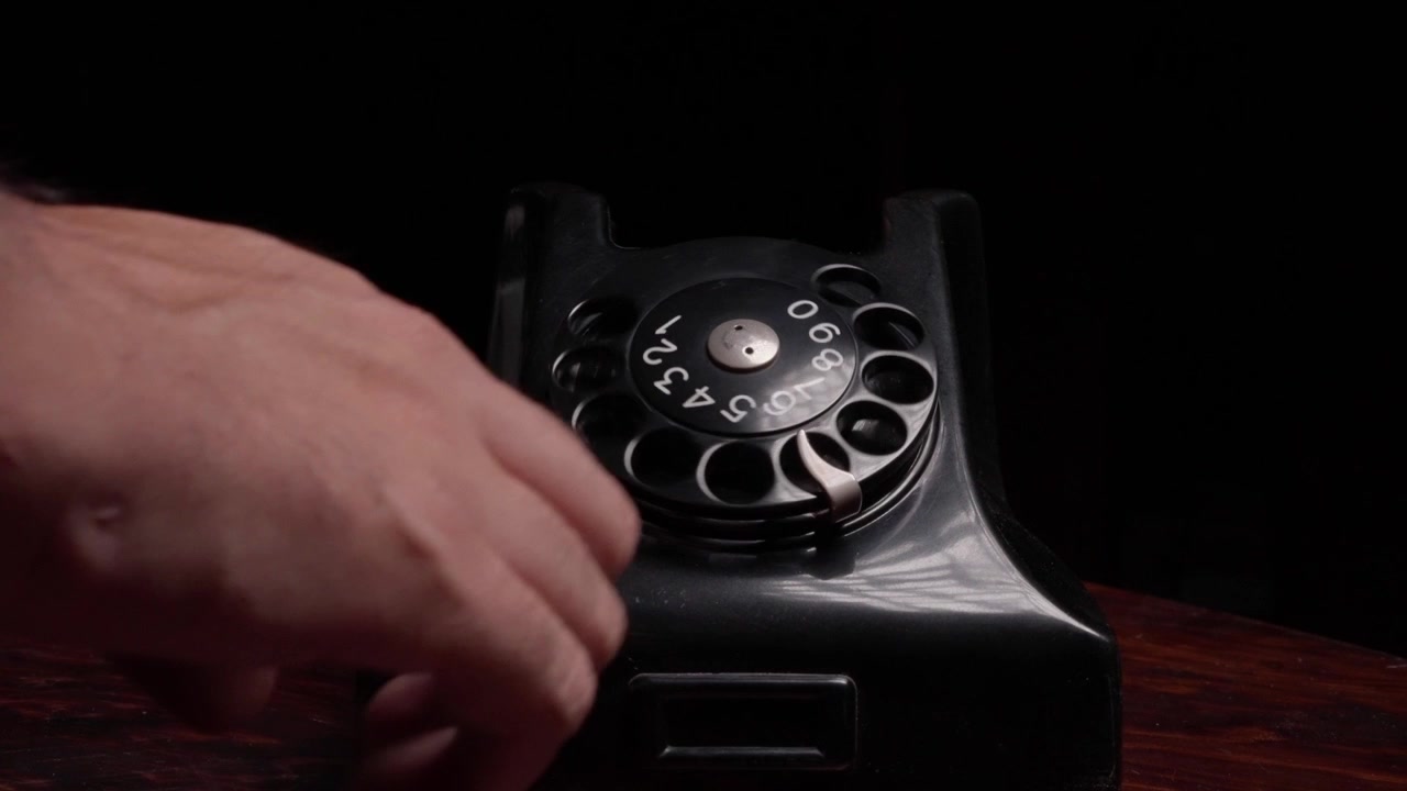 Tangan memegang gagang telepon di atas telepon putar vintage hitam di atas meja kayu, tampilan statis frontal medium saat mereka memutar nomor lalu meletakkan telepon dan menutupnya
