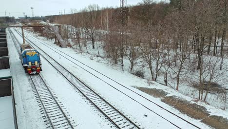 A freight train is passing by rail along rows of containers outside the city in the station area in the winter. Aerial view.