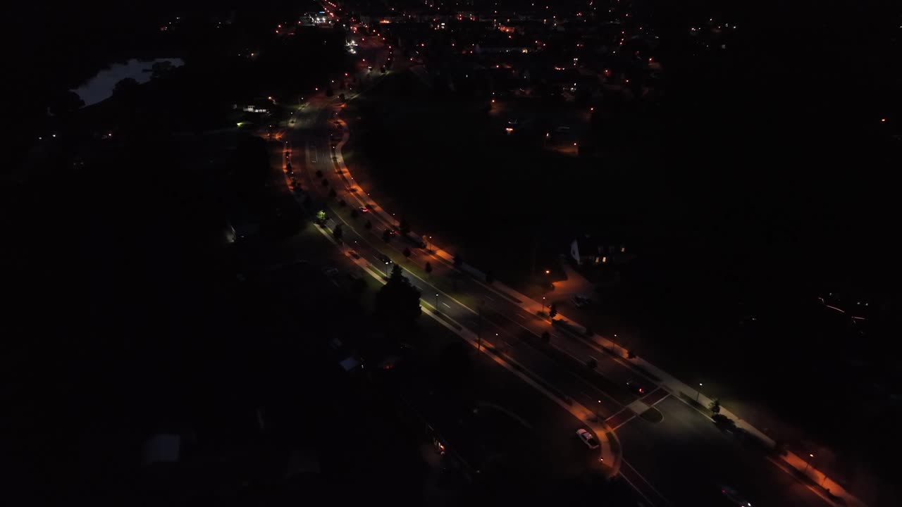 Night scene with warm glowing lantern along intersection road with traffic. Aerial flyover wide shot. Suburb neighborhood at midnight with driving cars. Illuminated cityscape in distance