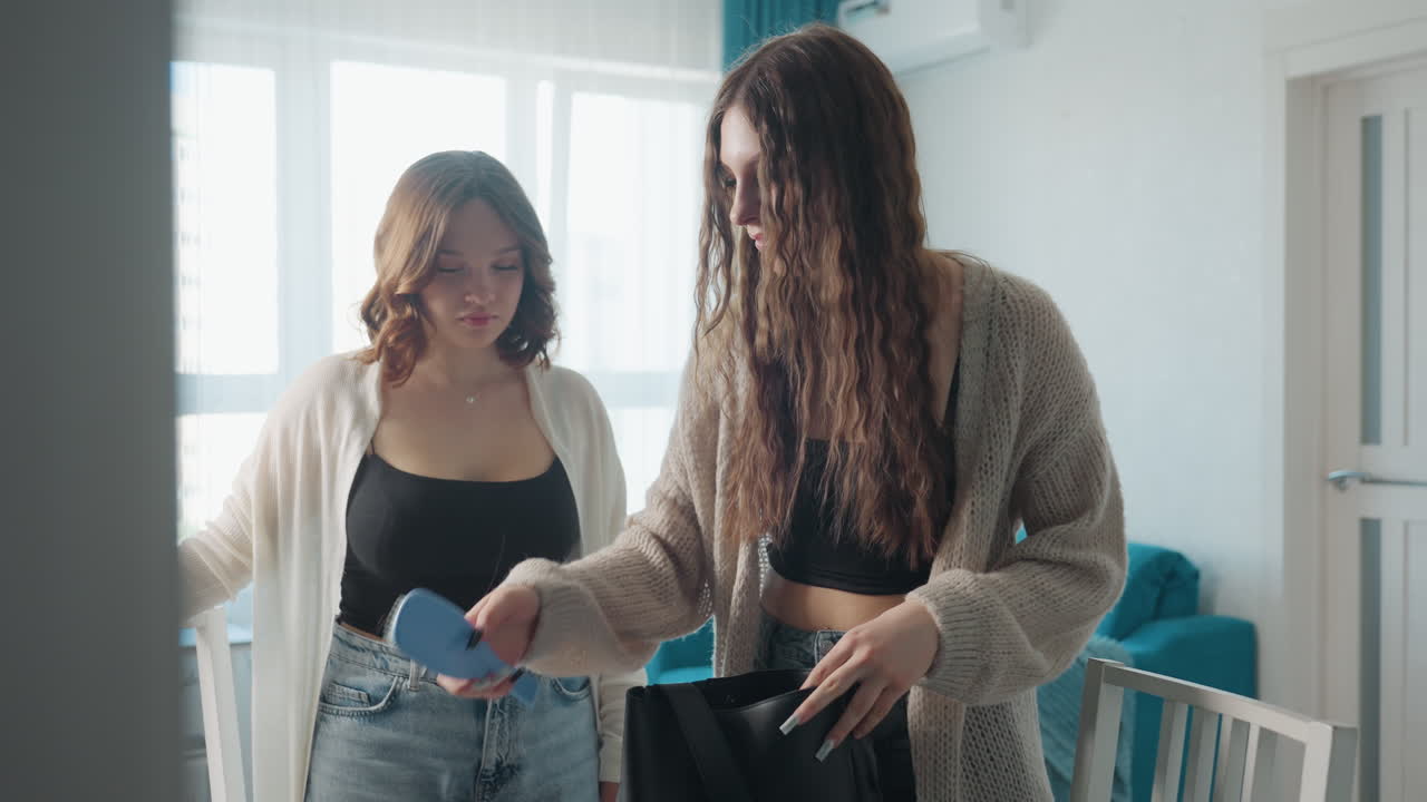 White Roommates Unpacking Purse At Table, Natural Morning Light Through Window, Two Young Women Examining Contents Of Black Bag, Denim Jeans And Cozy Cardigan, Relaxed Conversation And Friendly Smiles