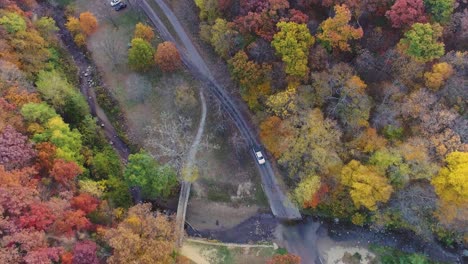 Aerial-tracking-shot-of-white-car-driving-through-autumn-forest-road-at-Ledges-State-Park