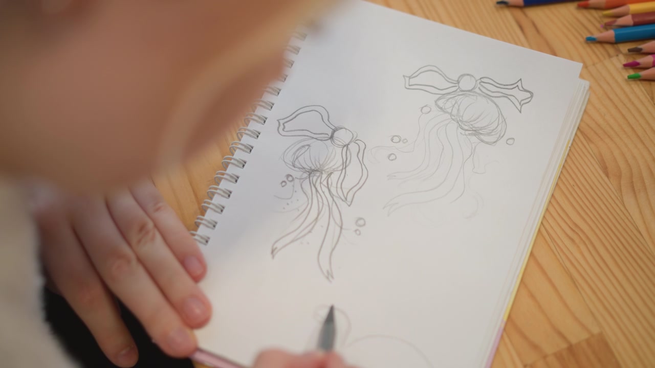 Vista ravvicinata da sopra la spalla mostra una persona che disegna una medusa e una piccola creatura su un quaderno a spirale, matita che si muove sulla carta su un tavolo di legno sotto una calda luce naturale, con schizzi e disegno.