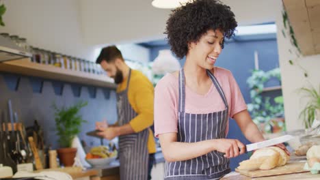 Video-of-happy-diverse-couple-in-aprons-baking-in-kitchen,-woman-slicing-bread,-with-copy-space