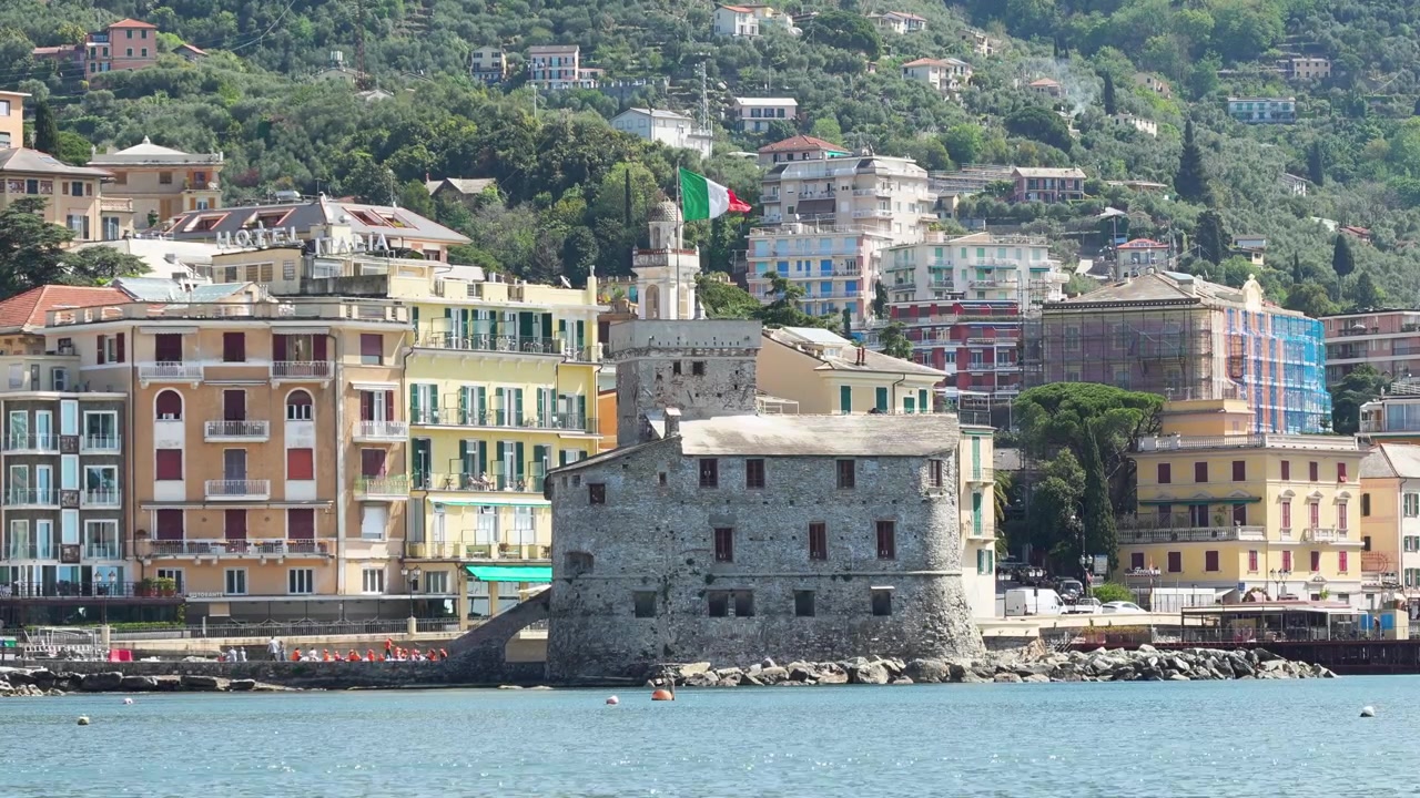 Scatto grandangolare diurno dello storico Castello di Rapallo sulla costa ligure, Italia. Vivaci edifici sul mare, bandiera italiana, colline lussureggianti e acqua azzurra evocano il fascino mediterraneo. Nessuna persona, statico.