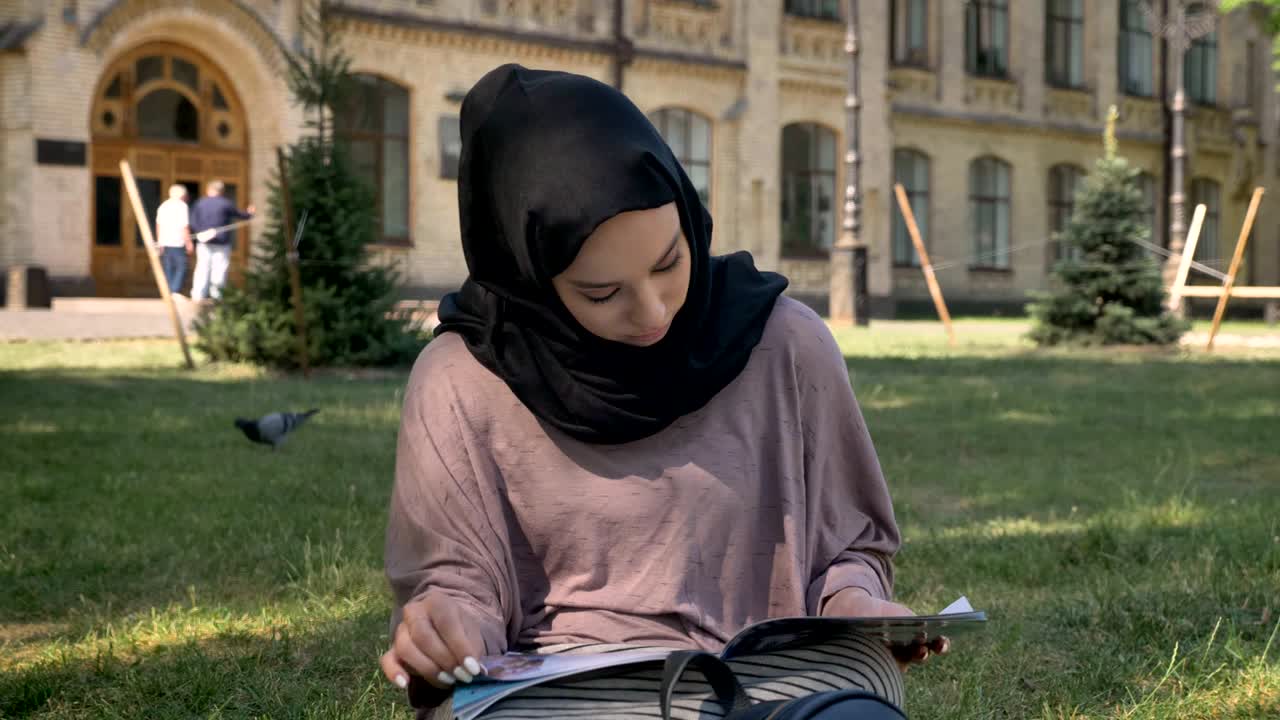 Young muslim girl in hijab is sitting on lawn and reading magazine, builging on background, religious concept, relax concept