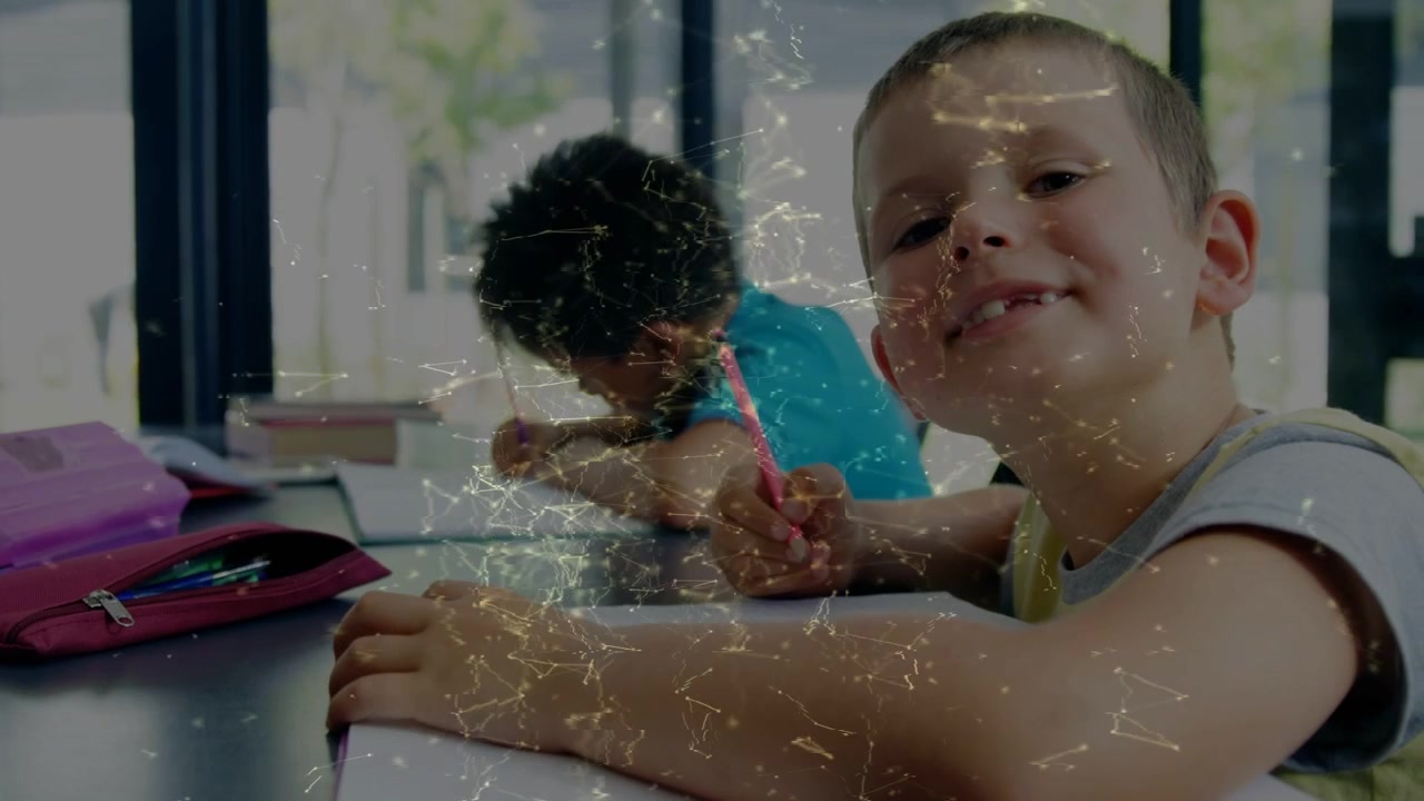 Alunos escrevendo, com o menino da frente levantando a cabeça e sorrindo para a câmera, segurando um lápis e partículas pela cena.