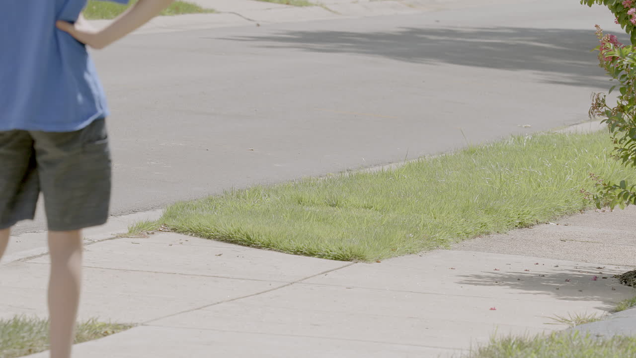 Medium shot of a young boy skating away from camera and down the sidewalk on a hover board in the suburbs on a sunny day in slow motion