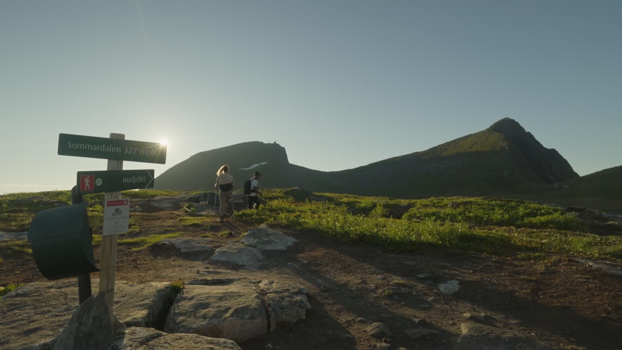pareja caminando en senja, noruega, bajo el sol de medianoche, pasando por una señal de sendero y usando palos