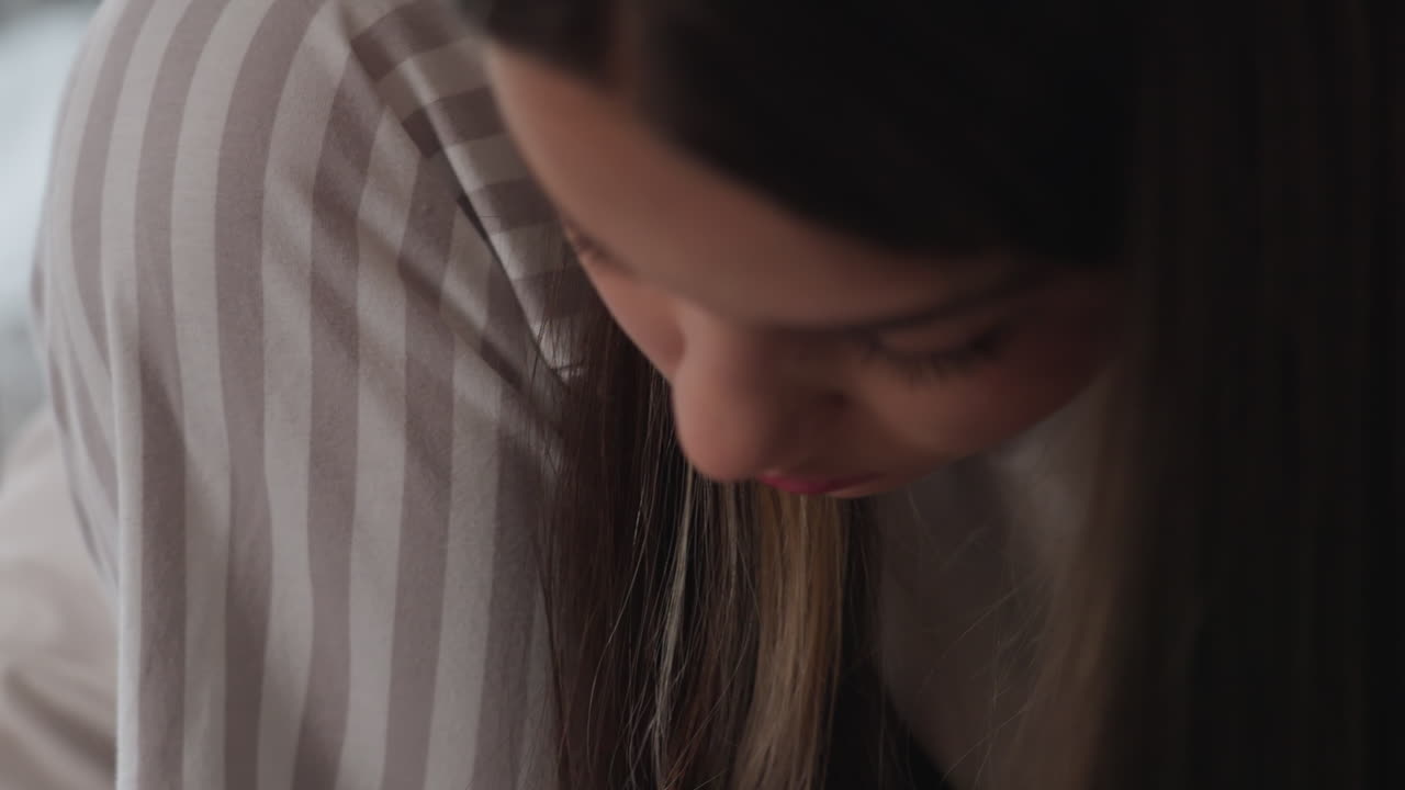 Side view female student with head bent painting toes with black nail polish, focused expression, soft indoor light, blurred rail background, striped trousers and white sleeve visible