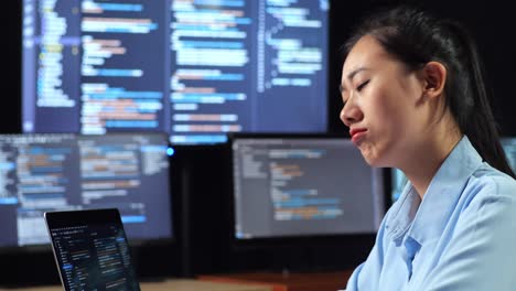 close up side view of asian female programmer thinking about something while writing code by a laptop using multiple monitors showing database on terminal window desktops in the office