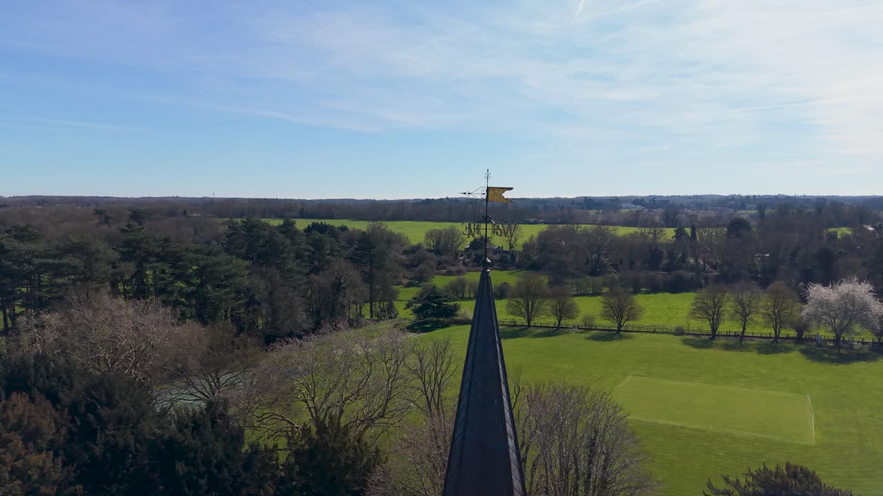 St Mary's Church spire and weather vane filmed by drone orbit in Hertingfordbury, Hertfordshire, England, with rural countryside, treetops and distant village under blue sky