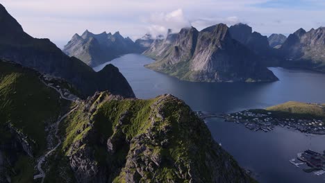 reinebringen mountain ridge summit with trail walkways - aerial orbiting motion, distant view of reine village at the mount base and lofoten islands surrounded by still water lakes and fjords