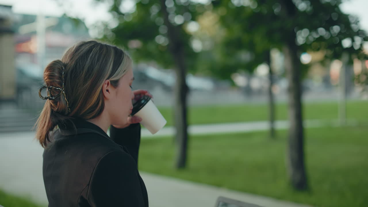 Side view of woman sipping beverage from paper cup while working on laptop seated in park with green grass and trees around her, calm urban background visible with soft light