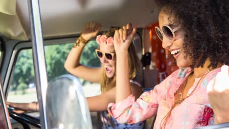 Two-diverse-female-friends-wearing-sunglasses-sitting-in-car-and-dancing