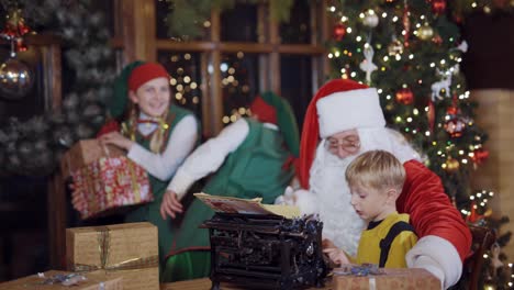 santa is teaching little boy to print on an old typing machine. christmas time. two elves in green costumes with presents in boxes.
