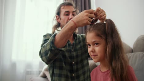 Un-Chico-Moreno-De-Pelo-Largo-Con-Una-Camisa-A-Cuadros-Verde-Le-Hace-A-Su-Pequeña-Hija-Con-Una-Chaqueta-Rosa-Un-Peinado-De-Cola-De-Caballo-Usando.-Un-Padre-Soltero-Moreno-Peina-A-Su-Hija-Con-Una-Banda-Elástica-Especial-En-Una-Casa-Moderna-Sentado-En-El-Sofá.