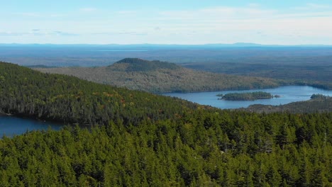 aerial drone shot flying over a forested ridge to reveal blue mountain lakes in the forest along a mountain range as summer ends and the season changes to fall in maine