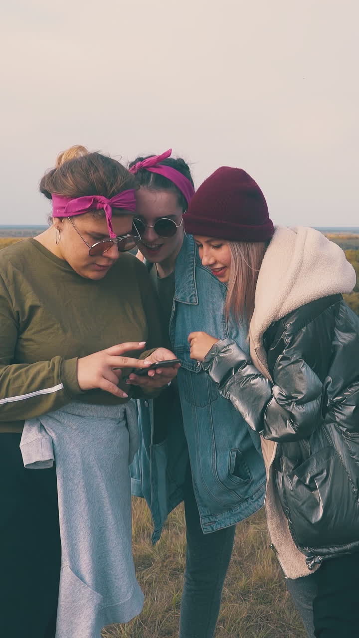 happy girls tourists choose music with mobile phone on hilly river edge against pictorial autumn landscape at sunset
