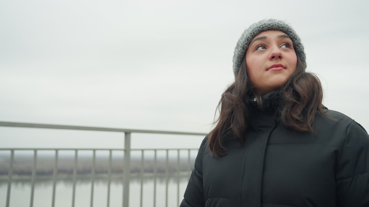 Woman looking up at overcast sky, closeup portrait in knit hat and puffer coat, soft smile with waterfront bridge and railing behind, expansive sky and quiet hopeful expression, urban overlook path