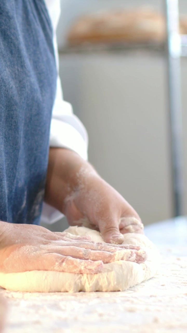 Vertical video: Lifting dough, baker in apron folding pressing on floury bakery bench for rising
