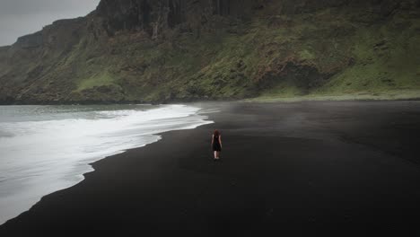 Young-beautiful-redhead-woman-walking-along-a-black-sand-beach-barefoot-in-a-black-dress
