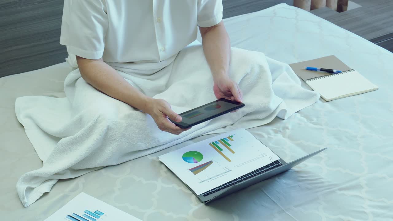 Asiatisk forretningsmand arbejder med tablet over papirdiagram eller graf i sengen