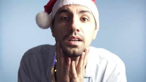 Closeup-footage-of-young-attractive-man-in-santa-claus-red-hat-after-celebration-or-long-night-looking-at-camera-mirror-and-touching-his-face,-recognizing-himself,-preen.-Background-isolated-on-blue