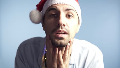 imágenes de primer plano de un joven atractivo con sombrero rojo de papá noel después de una celebración o una larga noche mirando el espejo de la cámara y tocándose la cara, reconociéndose a sí mismo, preen. fondo aislado en azul