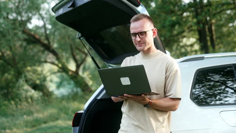 positive caucasian man works on his laptop. man stands near his car in the summer wood.