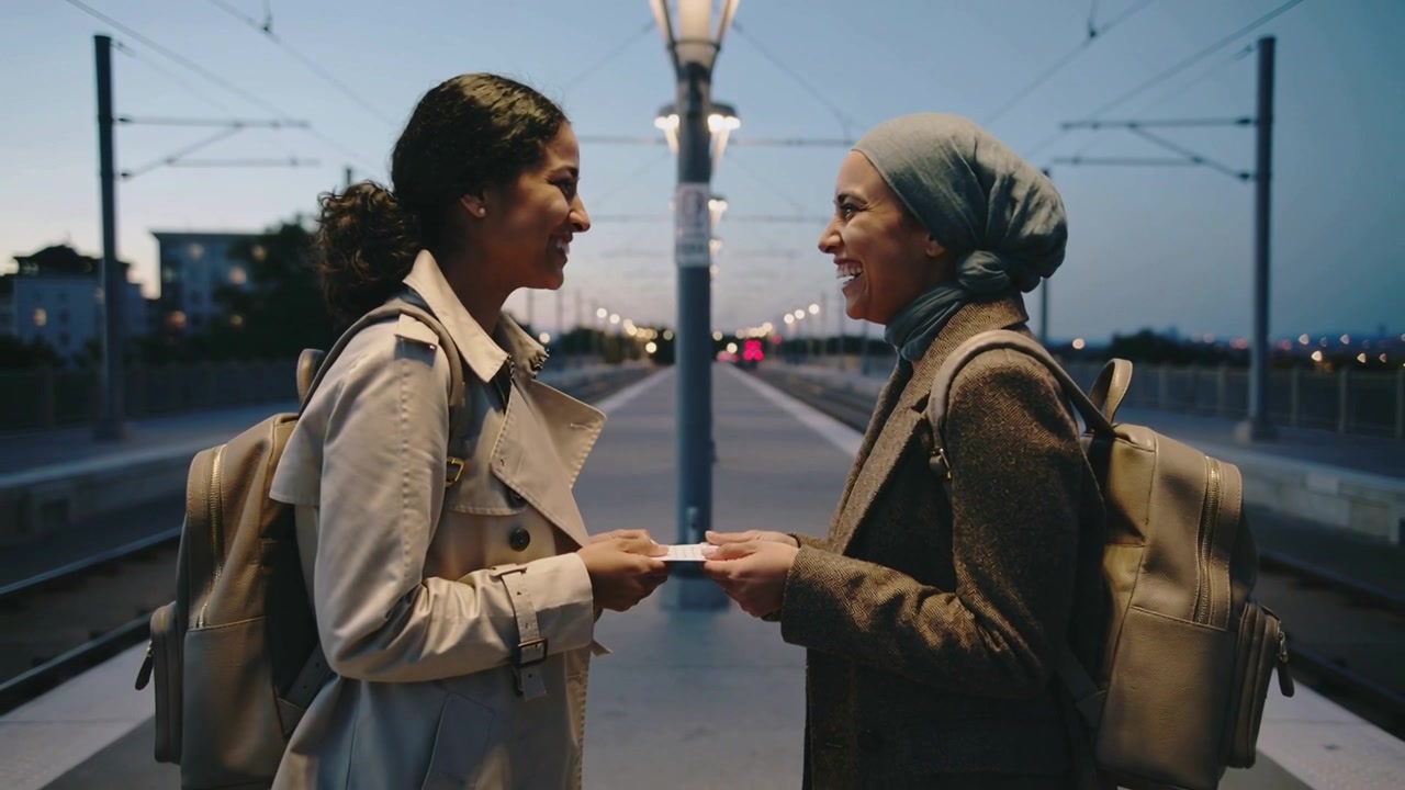 Bertemu di peron trem, dua wanita ber ransel dan berkerudung memegang kartu, tersenyum dan berpapasan