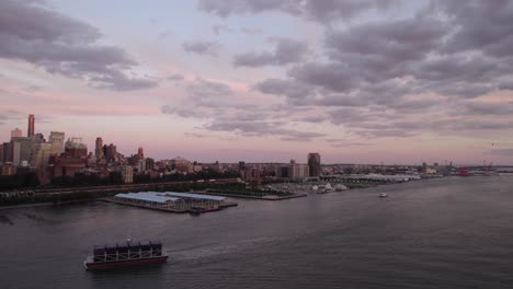 Aerial-view-of-a-container-ship-in-front-of-the-Brooklyn-cityscape,-colorful-dusk-in-NYC,-USA