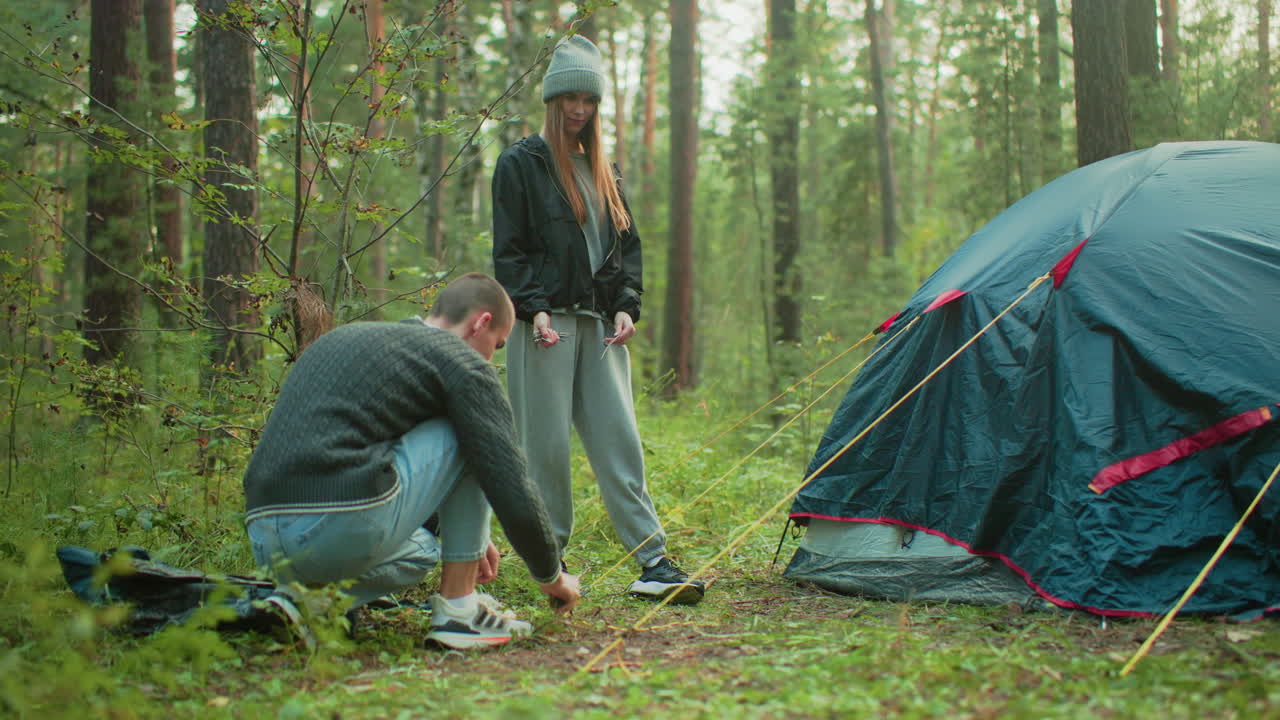 Woman in casual outfit stands nearby and watches as man crouches to peg yellow rope to forest ground beside dome tent during outdoor camping setup among trees in natural woodland setting