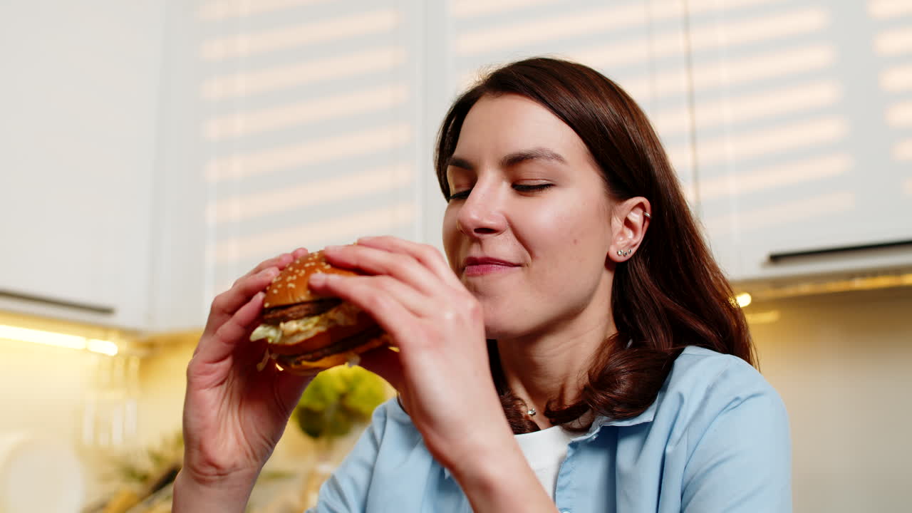 Young woman enjoying tasty hot burger fast food in home kitchen feeling happy satisfied during lunch