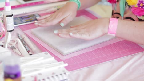 hands creating a scrapbook on pink mat with crafting tools around, bright colors