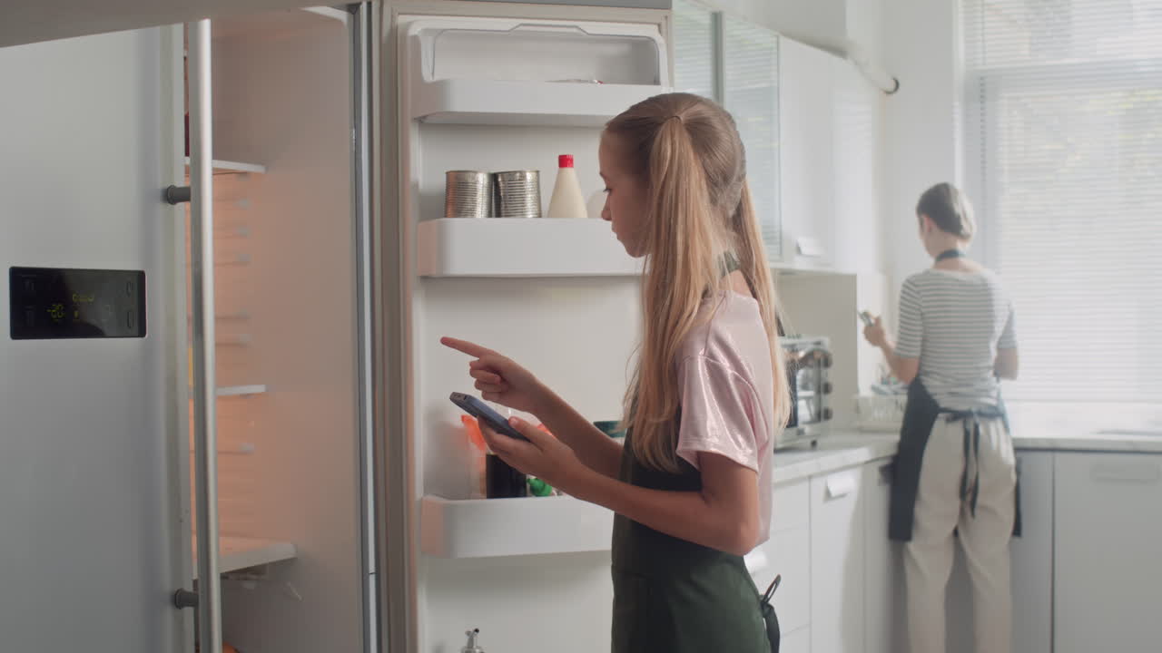 Girl Using List of Ingredients on Phone to Take Products from Fridge