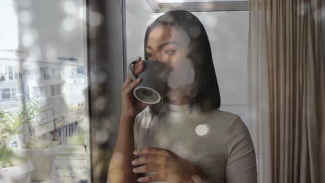 Animation-of-light-spots-over-biracial-woman-holding-mug-and-drinking