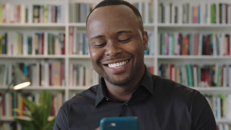 young-african-american-man-portrait-using-smartphone-laughing-standing-in-library-social-media
