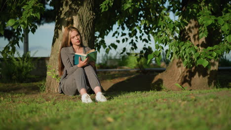 woman sitting outdoors, head slightly tilted, leaning against tree on grassy field, reading a book under warm sunlight, tree leaves sway gently in the breeze, background features greenery