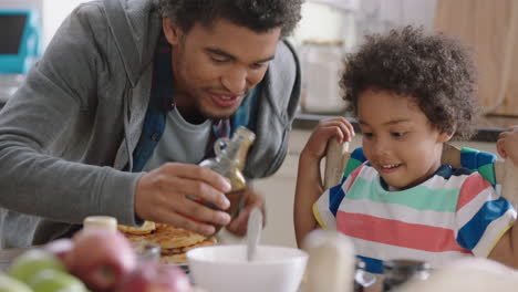 Fröhlicher-Kleiner-Junge,-Der-Zum-Frühstück-Frische-Waffeln-Isst,-Während-Sein-Vater-Honig-Einschenkt-Und-Köstliche-Hausgemachte-Leckereien-Für-Seinen-Sohn-In-Der-Heimischen-Küche-Zubereitet-4k