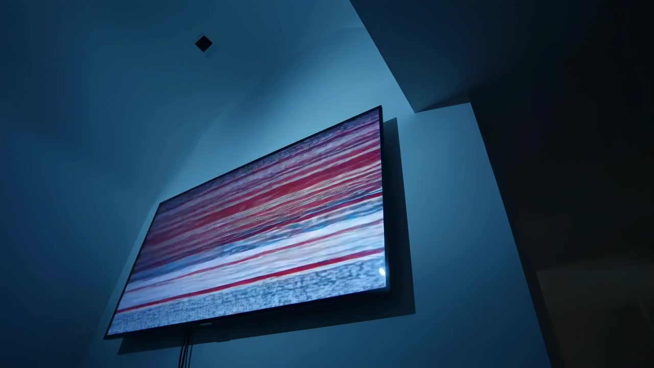 Mid frame in a dark interior shows a large blue illuminated wall mounted screen
