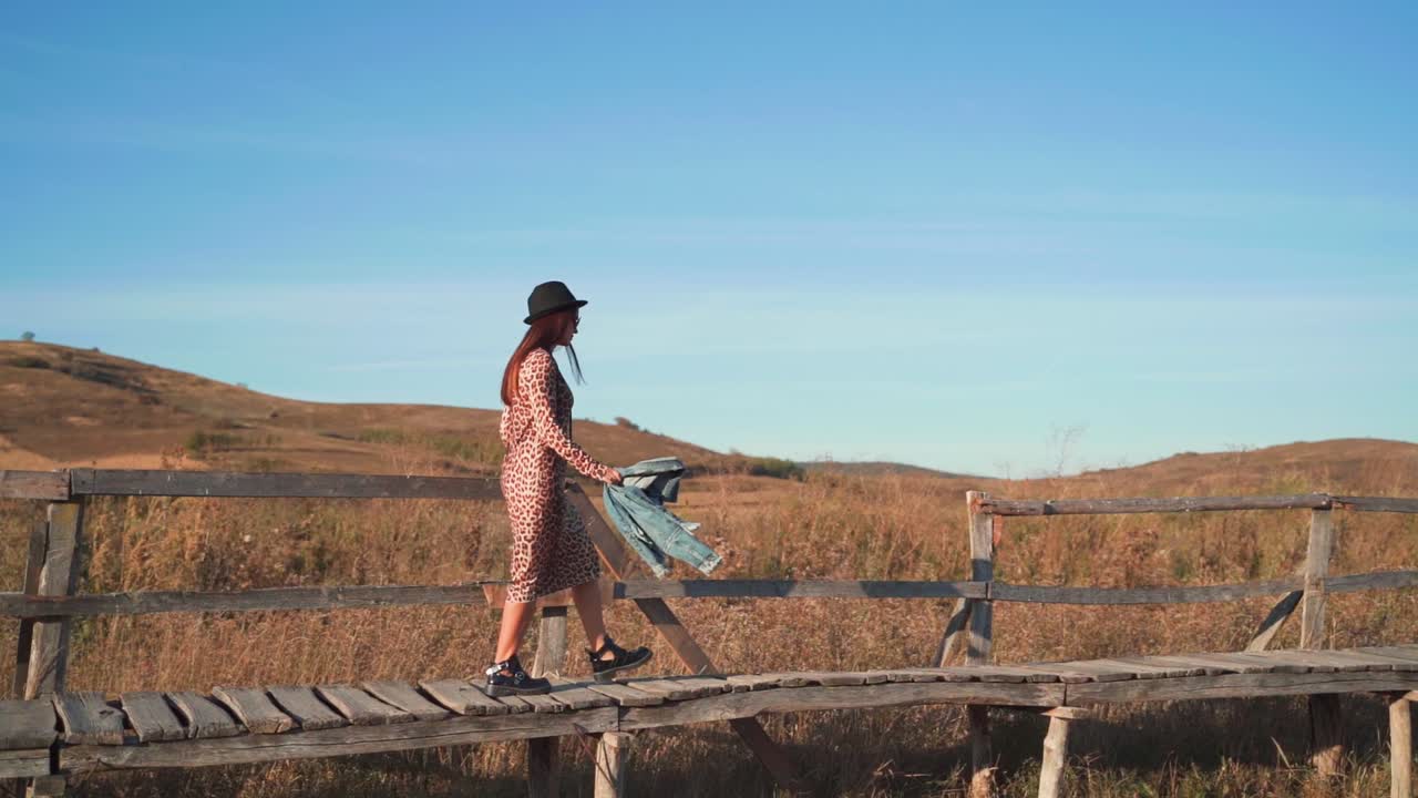 Young Caucasian woman wearing black hat, black shoes and printed animal dress carrying jean jacket walks on wooden boardwalk, handheld slow motion