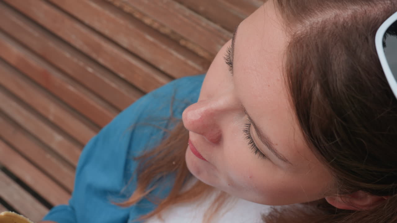 나무 벤치에 앉아 햇빛 아래 차분한 표정으로 웰빙을 나타내듯 생 플랜테인을 먹고 있는 여성의 클로즈업 오버헤드 샷. 자연스러운 질감이 강조된 평화로운 여름날의 한때