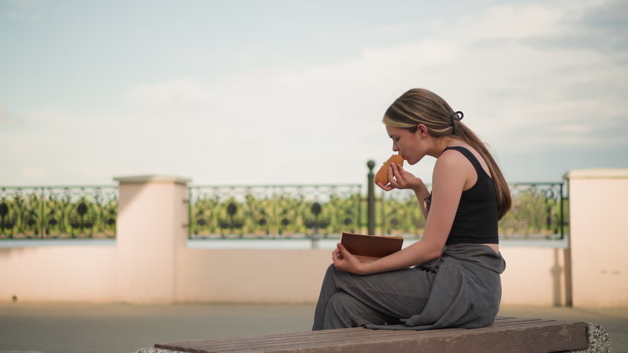 야외에 앉아있는 검은 크 을 입은 여성, 장식 울타리와 밝은 하늘을 배경으로 책을 읽는 동안 햄버거를 먹고, 야외 환경에서 읽고 간식을 먹습니다.